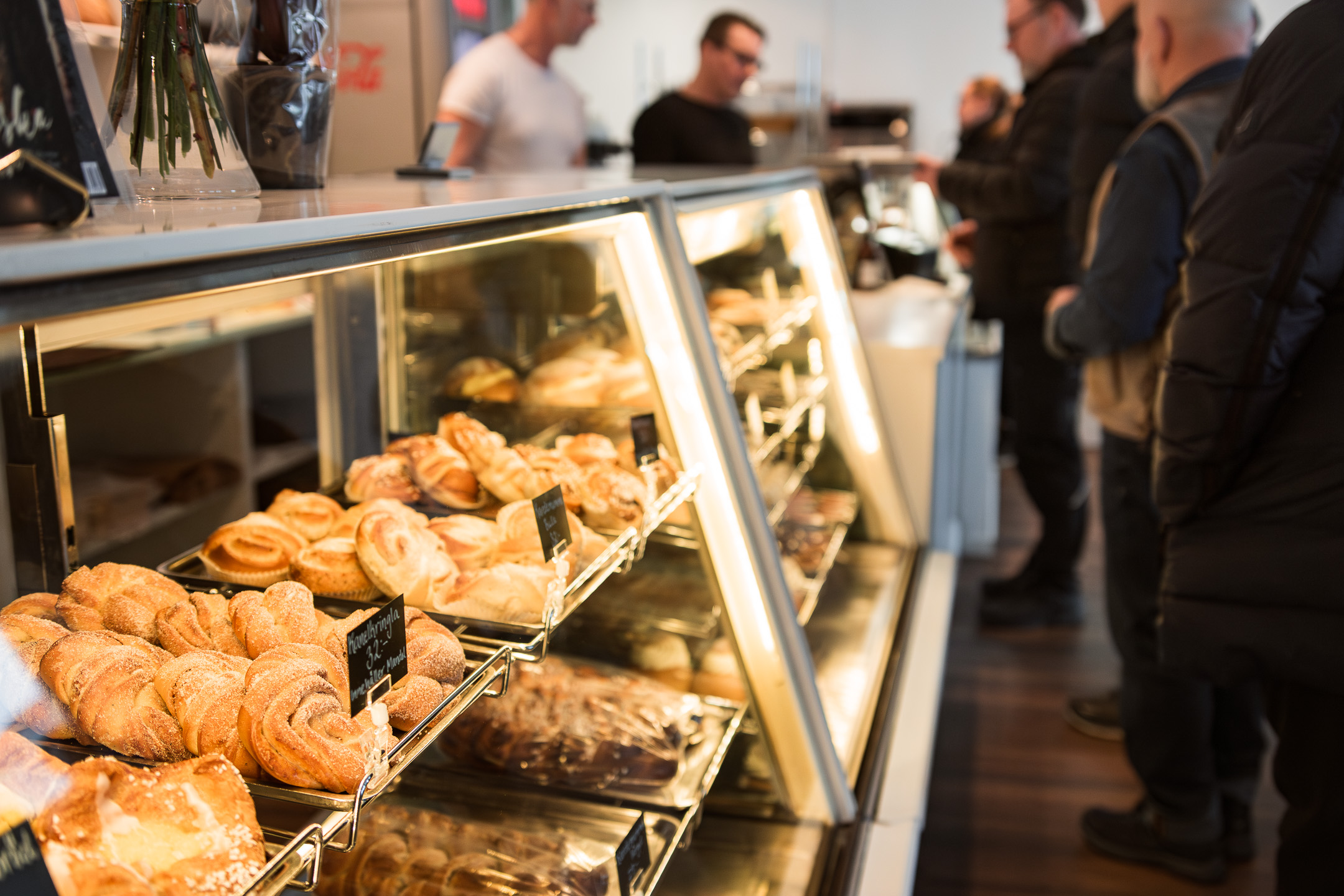 Kunder köar vid en cafédisk med en glasmonter fylld av färska bullar och bakverk.